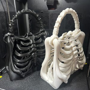 May include: Two 3D-printed skeleton-shaped handbags. One is black, the other is white. The bags have a handle and are designed to resemble a rib cage. The bags are sitting on a black surface.
