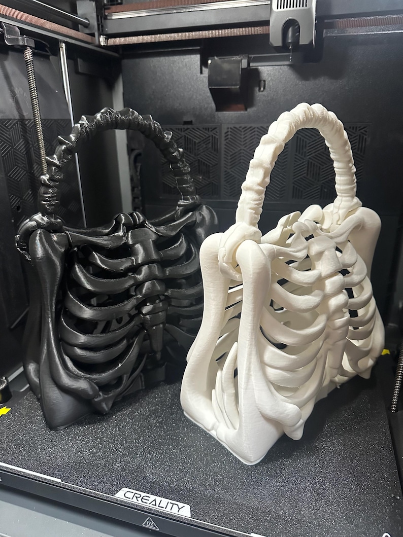 May include: Two 3D-printed skeleton-shaped handbags. One is black, the other is white. The bags have a handle and are designed to resemble a rib cage. The bags are sitting on a black surface.