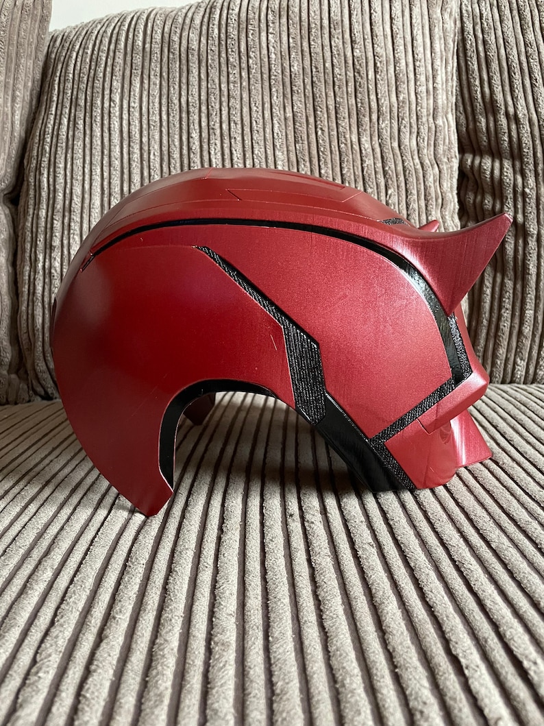 May include: A red and black Daredevil helmet with a textured surface. The helmet is made of plastic and has a sleek, modern design.