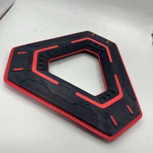 May include: A black and red geometric object with a textured surface. It features a hexagonal inner shape and a rectangular outer shape. Red accents highlight the edges and inner details. The object's dimensions are not visible.