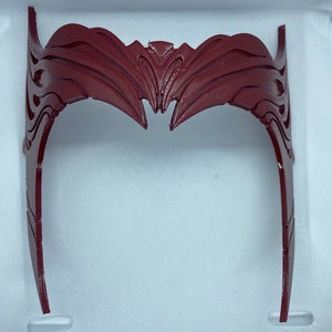 May include: A red plastic headband with a detailed, intricate design. The headband is shaped like a crown with a pointed top and curved sides.