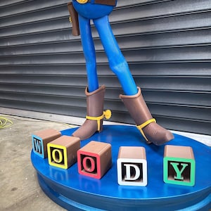 May include: A large statue of the Woody character from Toy Story. The statue has blue trousers, brown boots, and a yellow plaid shirt. The base is blue with blocks spelling out "WOODY".