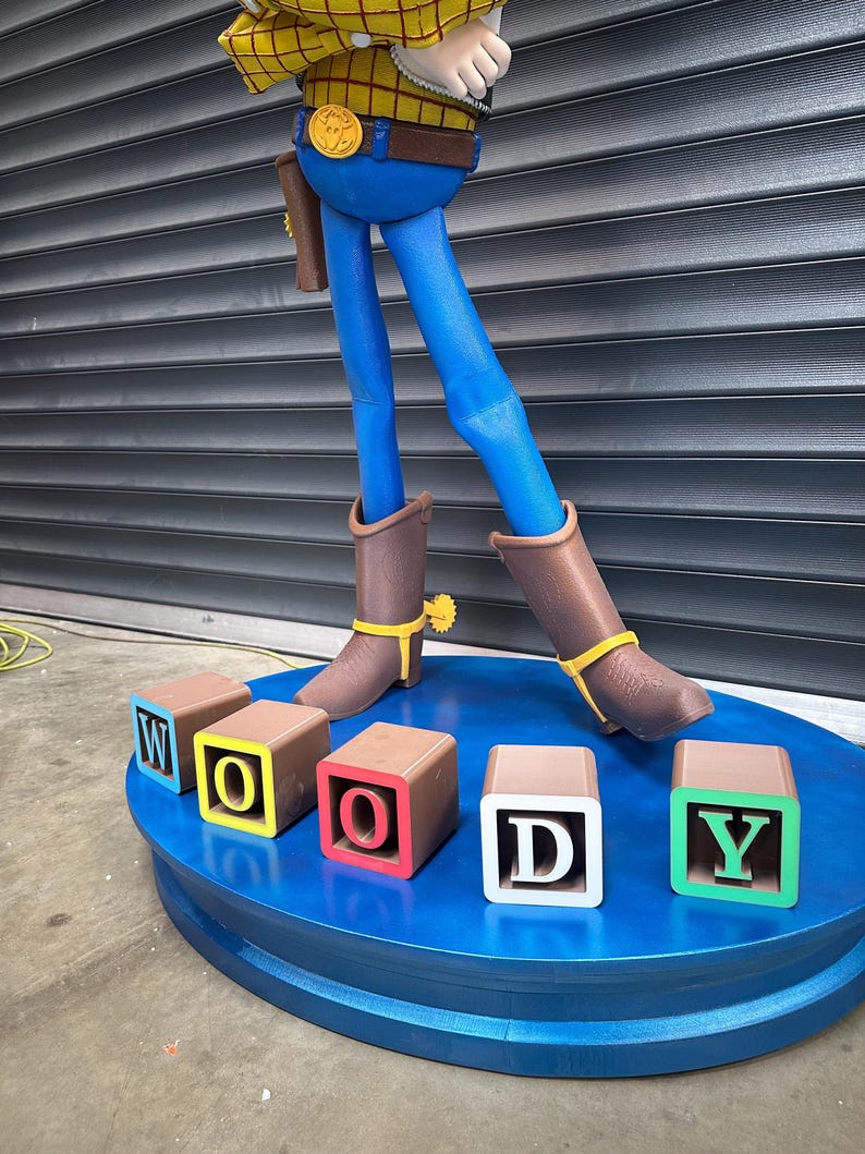 May include: A large statue of the Woody character from Toy Story. The statue has blue trousers, brown boots, and a yellow plaid shirt. The base is blue with blocks spelling out "WOODY".