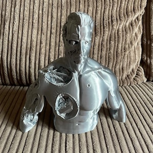 May include: A silver 3D printed bust of a man with a robotic, exposed chest. The bust is detailed with muscle definition and a realistic face.