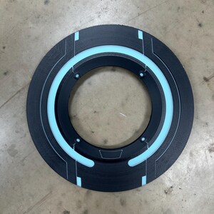 May include: A black circular object with a light blue glowing ring and accents. The object has a textured surface and a central opening. The design features geometric lines and shapes, creating a futuristic aesthetic.