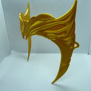 May include: A gold-coloured plastic crown with a detailed, intricate design. The crown is made of a single piece of plastic and has a curved shape.
