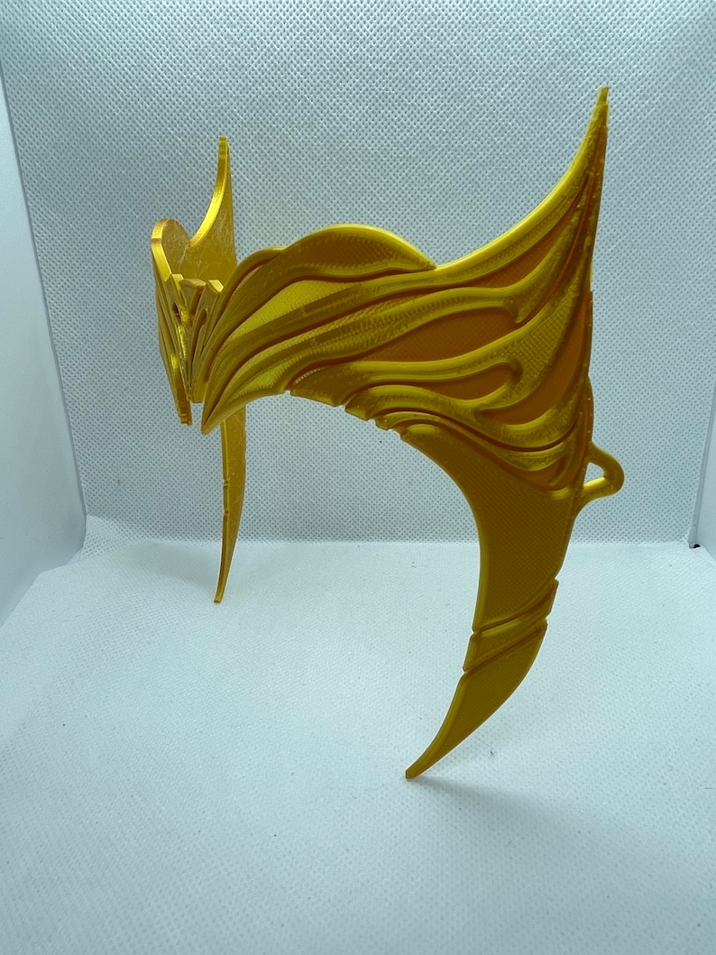May include: A gold-coloured plastic crown with a detailed, intricate design. The crown is made of a single piece of plastic and has a curved shape.