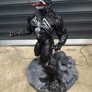 May include: A detailed black Venom statue with a white spider emblem on the chest. The figure has a long red tongue, sharp teeth, and is posed on a gray stone base. The statue is a collectible item.