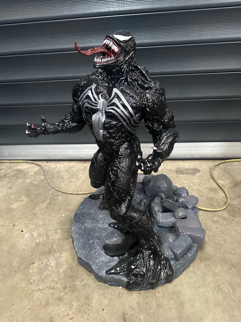 May include: A detailed black Venom statue with a white spider emblem on the chest. The figure has a long red tongue, sharp teeth, and is posed on a gray stone base. The statue is a collectible item.