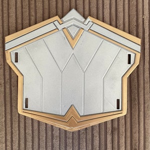May include: A silver and gold plastic shield with a geometric design. The shield is shaped like a heart with a pointed top.