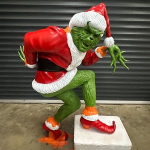 May include: A large, green Grinch statue wearing a red Santa suit with white trim and a matching hat. The Grinch is posed on a white and black box, with orange accents on his feet and shoes. The statue is a festive holiday decoration.