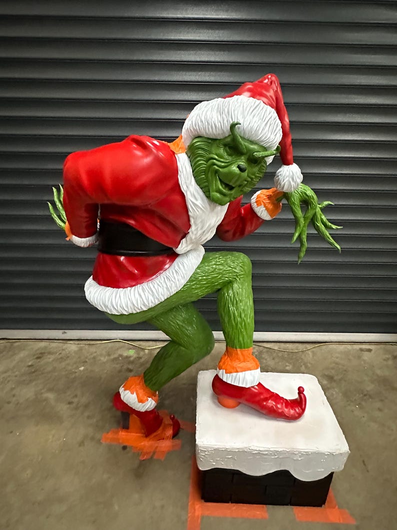 May include: A large, green Grinch statue wearing a red Santa suit with white trim and a matching hat. The Grinch is posed on a white and black box, with orange accents on his feet and shoes. The statue is a festive holiday decoration.