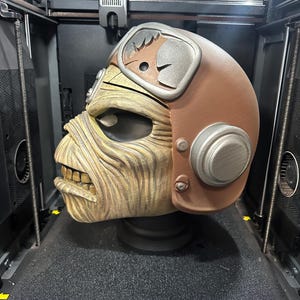 May include: A 3D-printed sculpture of a character's head, featuring a brown helmet with silver accents and a weathered, tan face. The sculpture is detailed and sits on a black base, inside a 3D printer.