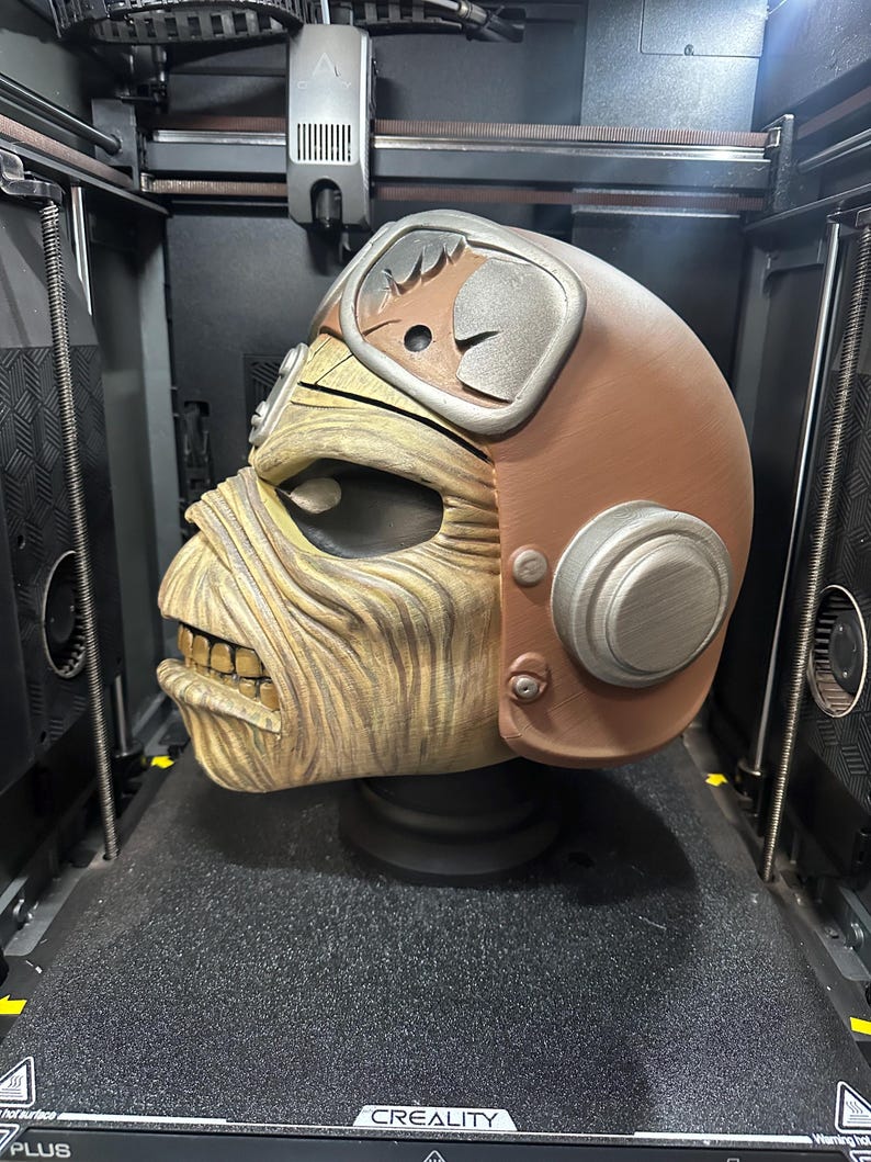 May include: A 3D-printed sculpture of a character's head, featuring a brown helmet with silver accents and a weathered, tan face. The sculpture is detailed and sits on a black base, inside a 3D printer.