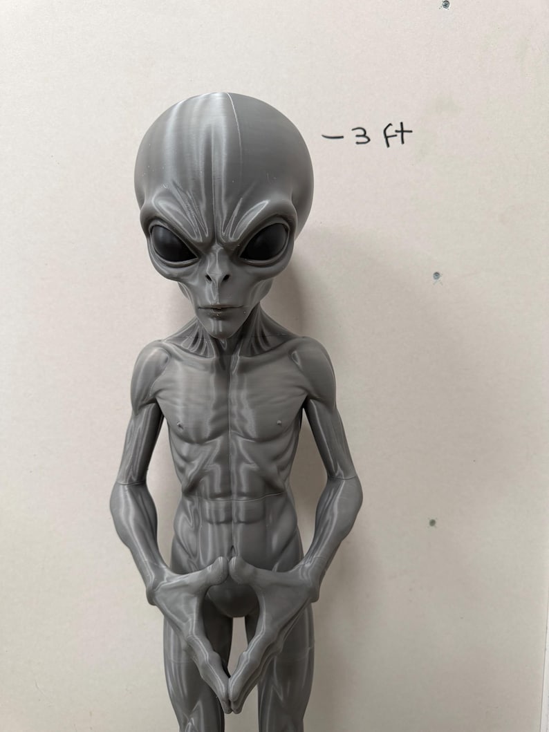 Pode incluir: Uma figura alien&iacute;gena cinza impressa em 3D com uma cabe&ccedil;a grande, lisa e alongada, olhos ovais escuros e um torso esguio e musculoso. A figura tem as m&atilde;os posicionadas juntas na frente da cintura. O texto - 3 ft est&aacute; escrito na parede.