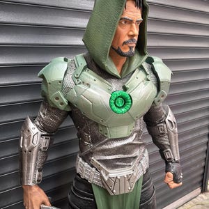 May include: A detailed statue of a character in a green hooded cloak and armor. The figure has a stern expression and a goatee. The armor is a mix of silver and green, with a glowing green chest piece. The figure is holding a silver gauntlet.