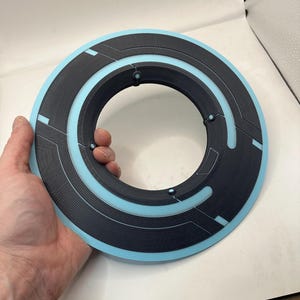 May include: A black and light blue circular object with a central hole. The object has a textured surface with geometric patterns and is held in a hand. The design features a light blue inner ring and accents.