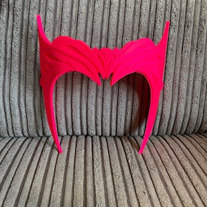 May include: A bright pink plastic headband with a detailed, intricate design. The headband is shaped like a crown with two points on the top.
