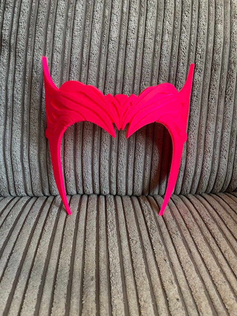 May include: A bright pink plastic headband with a detailed, intricate design. The headband is shaped like a crown with two points on the top.