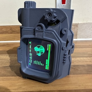 May include: A blue 3D printed replica of a Pip-Boy from the Fallout video game series. The screen displays a character's stats and special perks. The device has a rotary dial and buttons.