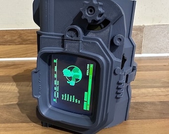 Könnte beinhalten: Eine blaue 3D-gedruckte Nachbildung eines Pip-Boys aus der Fallout-Videospielserie. Der Bildschirm zeigt die Statistiken und Spezialvorteile eines Charakters an. Das Gerät verfügt über einen Drehknopf und Tasten.