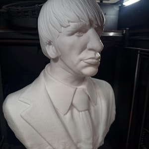 May include: A white bust sculpture of a man with short hair, a suit jacket, collared shirt, and tie. The sculpture is set against a dark background. The bust is a detailed representation of a person.