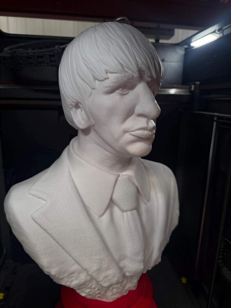 May include: A white bust sculpture of a man with short hair, a suit jacket, collared shirt, and tie. The sculpture is set against a dark background. The bust is a detailed representation of a person.