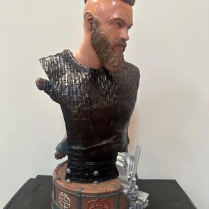 May include: A detailed bust of a man with a beard and shaved head, wearing a dark textured top. The bust is mounted on a round base with Celtic knot designs and a silver accent. The sculpture is a detailed representation.