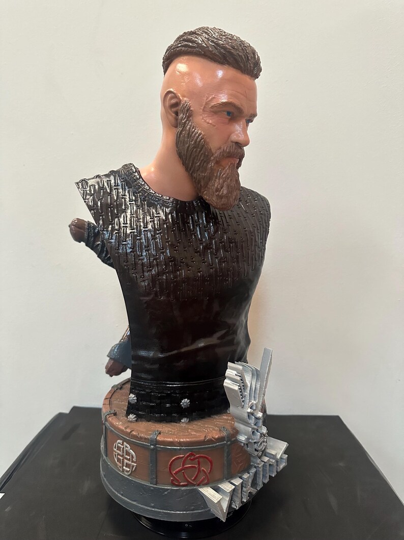 May include: A detailed bust of a man with a beard and shaved head, wearing a dark textured top. The bust is mounted on a round base with Celtic knot designs and a silver accent. The sculpture is a detailed representation.
