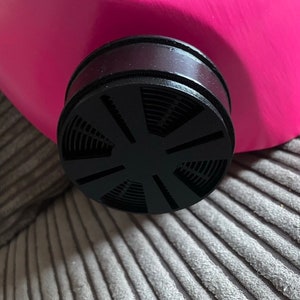 K&ouml;nnte beinhalten: Nahaufnahme eines schwarzen Gasmaskenfilters mit kreisf&ouml;rmigem Design und einer rosafarbenen Gasmaske im Hintergrund.