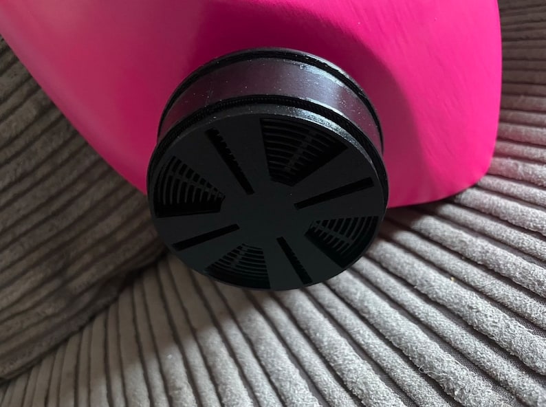 K&ouml;nnte beinhalten: Nahaufnahme eines schwarzen Gasmaskenfilters mit kreisf&ouml;rmigem Design und einer rosafarbenen Gasmaske im Hintergrund.