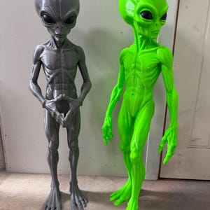 May include: Two standing alien figurines, one in metallic gray and one in neon green, both featuring large heads, dark almond-shaped eyes, and slender bodies. The gray figure has hands joined together, while the green figure stands with arms at its sides. Text reads -3 ft.