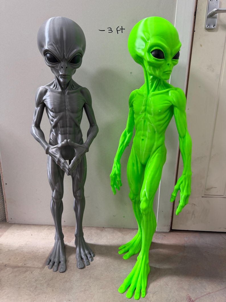 Pode incluir: Duas figuras alien&iacute;genas em p&eacute;, uma em cinza met&aacute;lico e outra em verde neon, ambas com cabe&ccedil;as grandes, olhos escuros em forma de am&ecirc;ndoa e corpos esguios. A figura cinza tem as m&atilde;os juntas, enquanto a verde tem os bra&ccedil;os ao lado do corpo. O texto diz -3 ft.