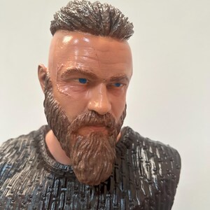 May include: A bust of a man with a brown beard and hair, and blue eyes. The figure has a scar on the face and is wearing a dark textured shirt. The bust is a detailed representation of a historical or fictional character.
