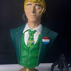 May include: A green bust of a character with a golden horned helmet, green suit, and a patterned tie. The bust has a gold base with the name "LOKI" in green. A button with the word "LOKI" is pinned on the suit.