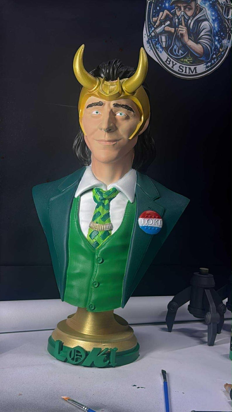May include: A green bust of a character with a golden horned helmet, green suit, and a patterned tie. The bust has a gold base with the name "LOKI" in green. A button with the word "LOKI" is pinned on the suit.