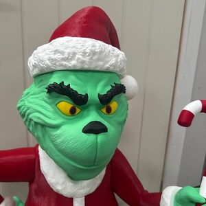 May include: A green character statue wearing a red Santa hat with white trim and a red suit. The character has yellow eyes, a black nose, and a grumpy expression. It is holding a candy cane.