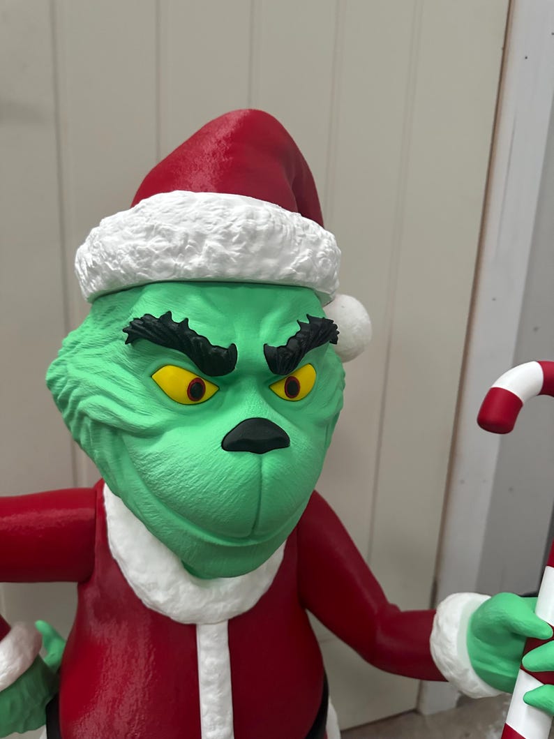 May include: A green character statue wearing a red Santa hat with white trim and a red suit. The character has yellow eyes, a black nose, and a grumpy expression. It is holding a candy cane.