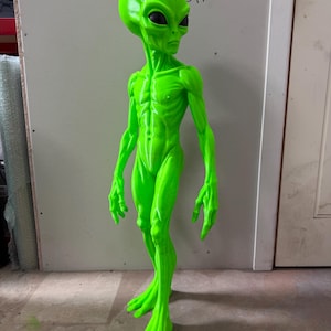 Pode incluir: Uma figura alien&iacute;gena verde n&eacute;on brilhante com uma cabe&ccedil;a alongada, grandes olhos pretos em forma de am&ecirc;ndoa e um corpo esguio e texturizado com musculatura definida. A figura est&aacute; de p&eacute; com bra&ccedil;os longos e m&atilde;os e p&eacute;s.