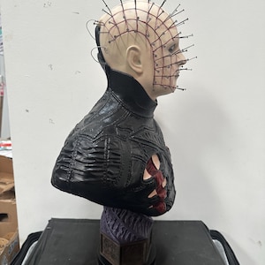May include: A bust of a character with a pale complexion, adorned with numerous metal pins embedded in the head. The figure wears a black, textured garment with a detailed design. The base is a dark purple color.