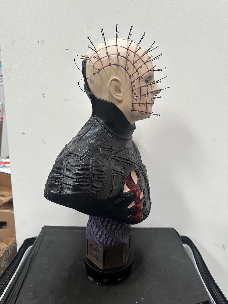 May include: A bust of a character with a pale complexion, adorned with numerous metal pins embedded in the head. The figure wears a black, textured garment with a detailed design. The base is a dark purple color.