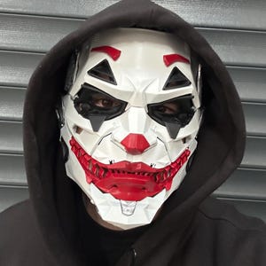 May include: A white and red Joker-style mask with a black hood. The mask has a geometric design with red lips and accents, and black eye openings. The hood is black.