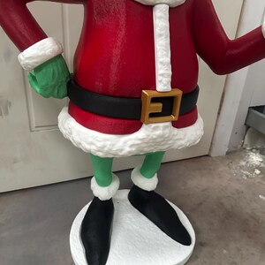 May include: A green character statue wearing a red coat with white trim, a black belt with a gold buckle, and black shoes. The statue has green hands and legs, and is standing on a white base. The statue is a Christmas decoration.