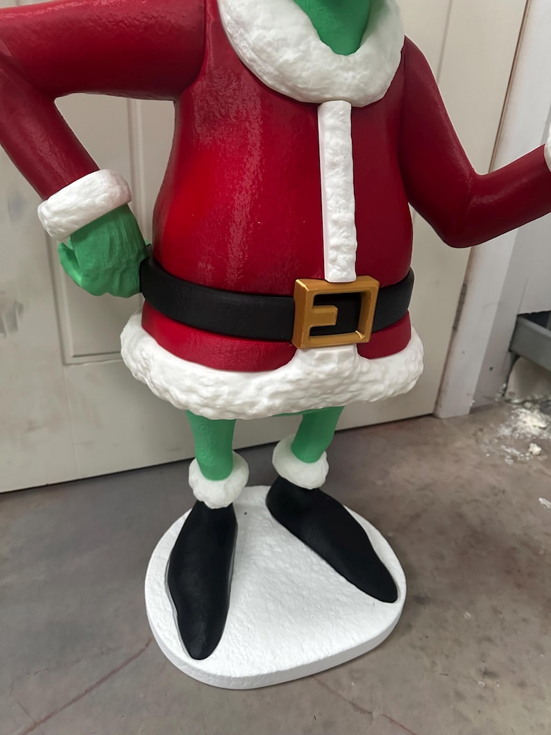 May include: A green character statue wearing a red coat with white trim, a black belt with a gold buckle, and black shoes. The statue has green hands and legs, and is standing on a white base. The statue is a Christmas decoration.