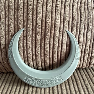 May include: A gray crescent-shaped object with a raised design of text and symbols around the edge.