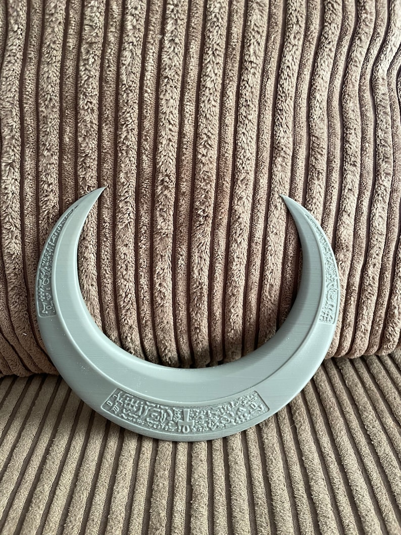 May include: A gray crescent-shaped object with a raised design of text and symbols around the edge.