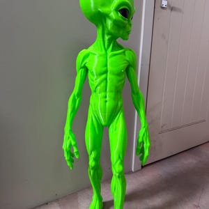 Pode incluir: Uma estatueta alien&iacute;gena em verde n&eacute;on brilhante com uma cabe&ccedil;a grande, lisa e de formato oval, com olhos escuros em forma de am&ecirc;ndoa. A figura tem um corpo humanoide esguio com defini&ccedil;&atilde;o muscular vis&iacute;vel, bra&ccedil;os longos e dedos alongados.