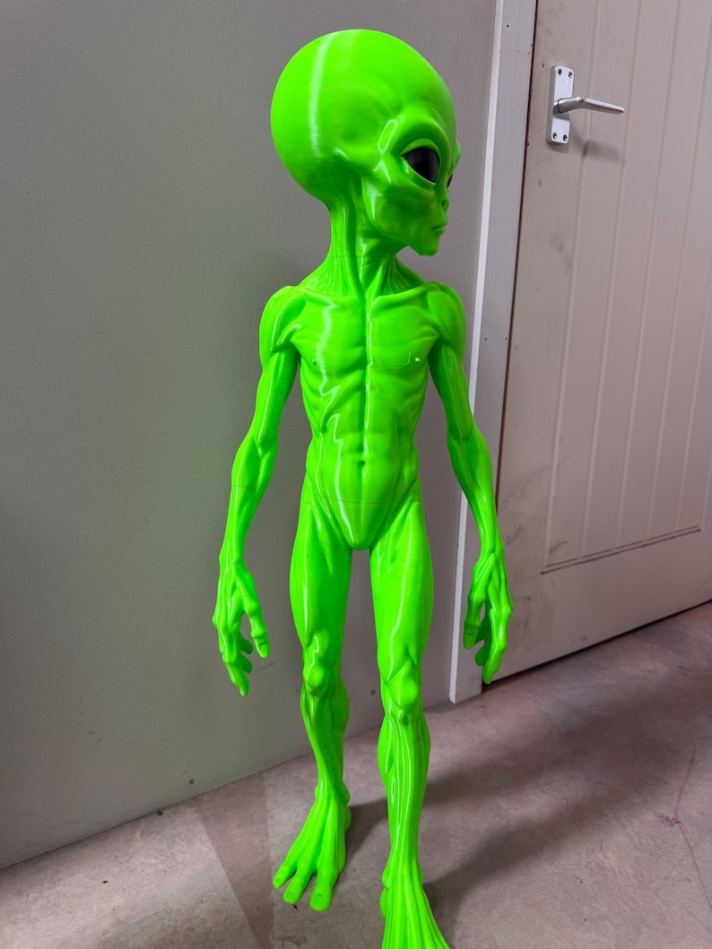 Pode incluir: Uma estatueta alien&iacute;gena em verde n&eacute;on brilhante com uma cabe&ccedil;a grande, lisa e de formato oval, com olhos escuros em forma de am&ecirc;ndoa. A figura tem um corpo humanoide esguio com defini&ccedil;&atilde;o muscular vis&iacute;vel, bra&ccedil;os longos e dedos alongados.