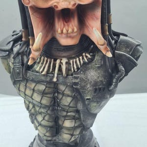 Puede incluir: Busto detallado de un alien&iacute;gena Predator, con una cara marr&oacute;n y beige texturizada, dientes afilados y rastas. La figura tiene una armadura de pecho detallada y una base, mostrando un tema de ciencia ficci&oacute;n.