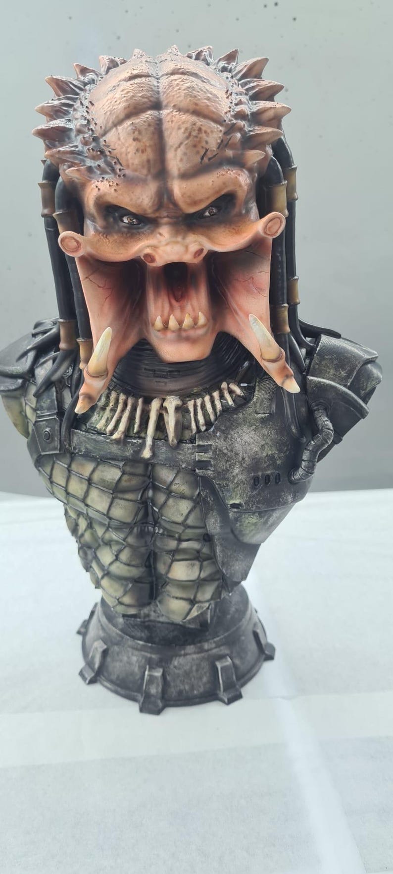 Puede incluir: Busto detallado de un alien&iacute;gena Predator, con una cara marr&oacute;n y beige texturizada, dientes afilados y rastas. La figura tiene una armadura de pecho detallada y una base, mostrando un tema de ciencia ficci&oacute;n.